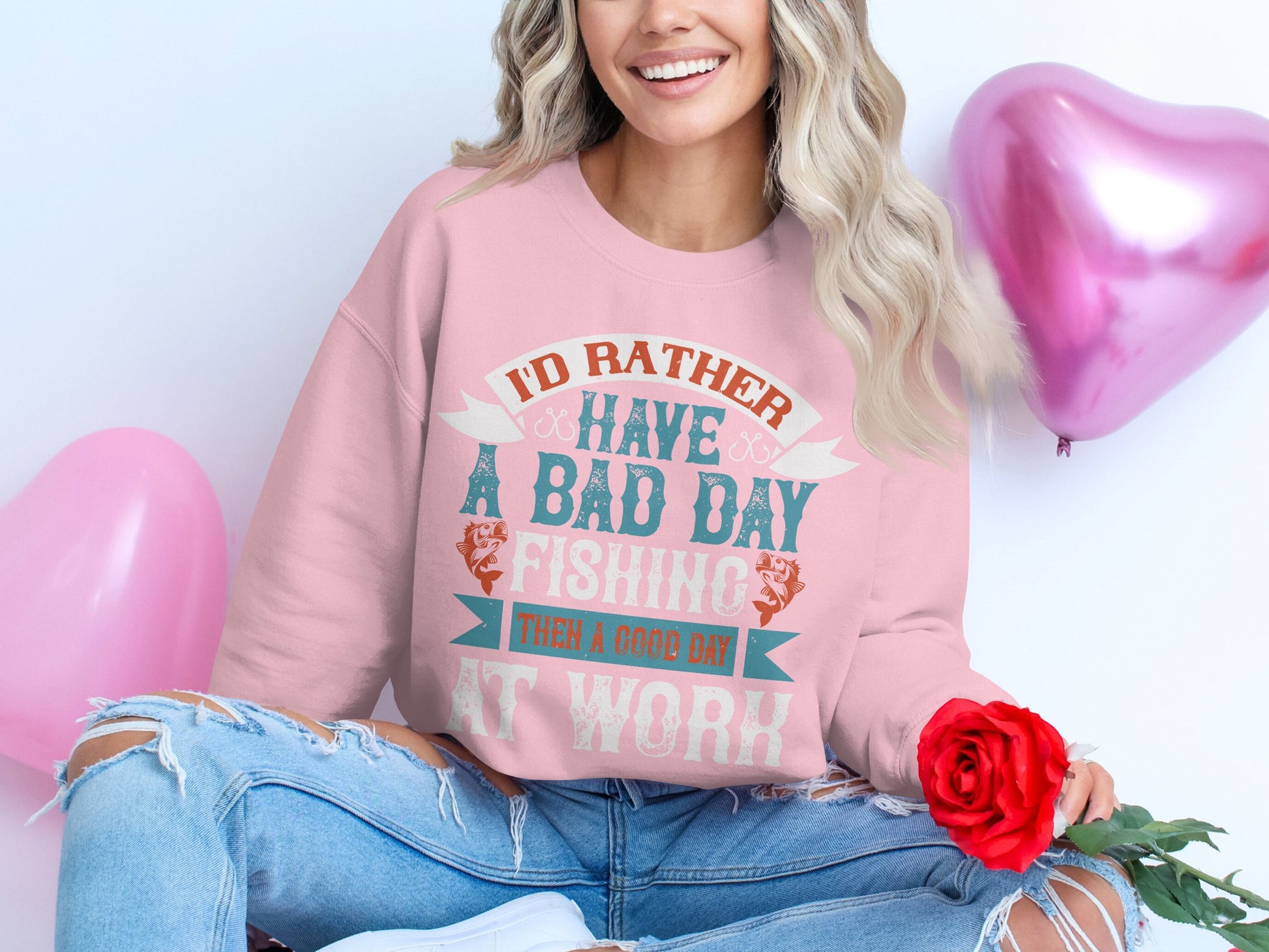 A person sits cross-legged on the floor wearing a pink Garment Graphics unisex fishing sweatshirt that says, Id rather have a bad day fishing than a good day at work. They hold a red rose as two pink heart-shaped balloons float in the background, highlighting their love for fishing.