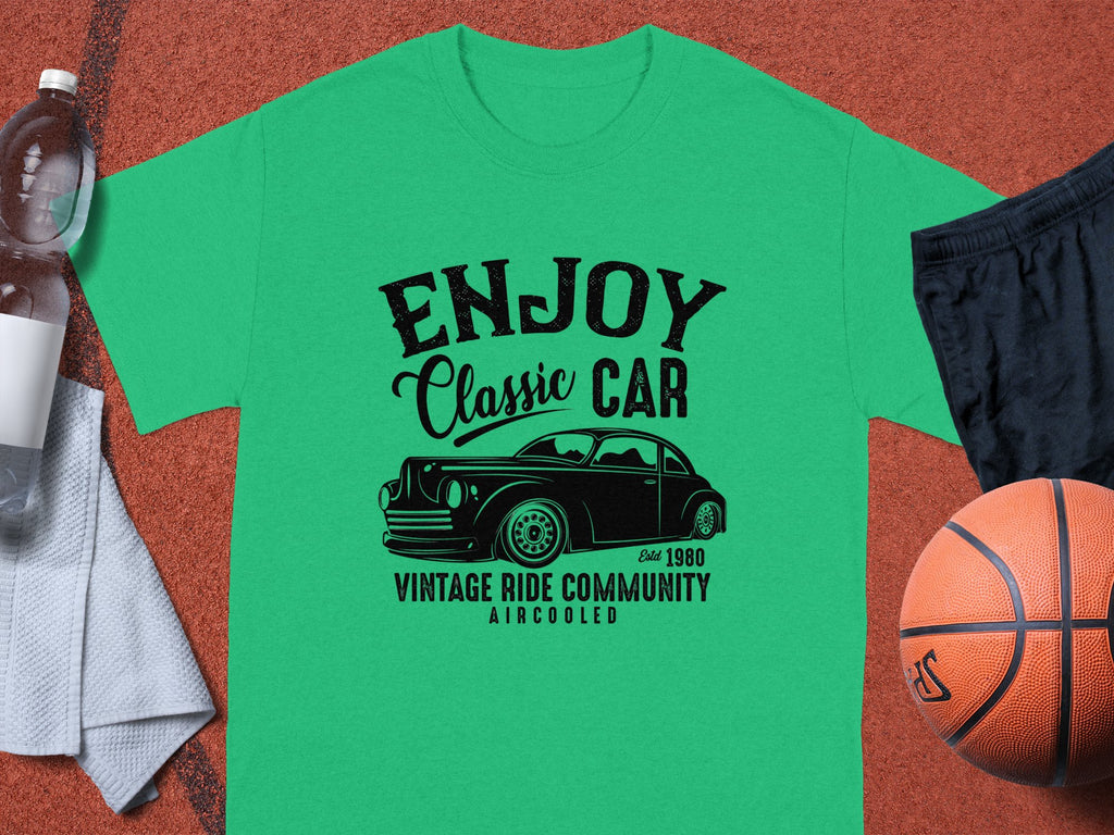 The Garment Graphics green t-shirt, ideal for car enthusiasts, showcases a Enjoy Classic Car design with a vintage car graphic and text Established 1980, Vintage Ride Community, Aircooled. Its displayed on a brown surface alongside a basketball, black shorts, towel, and water bottle.