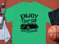 The Garment Graphics green t-shirt, ideal for car enthusiasts, showcases a Enjoy Classic Car design with a vintage car graphic and text Established 1980, Vintage Ride Community, Aircooled. Its displayed on a brown surface alongside a basketball, black shorts, towel, and water bottle.