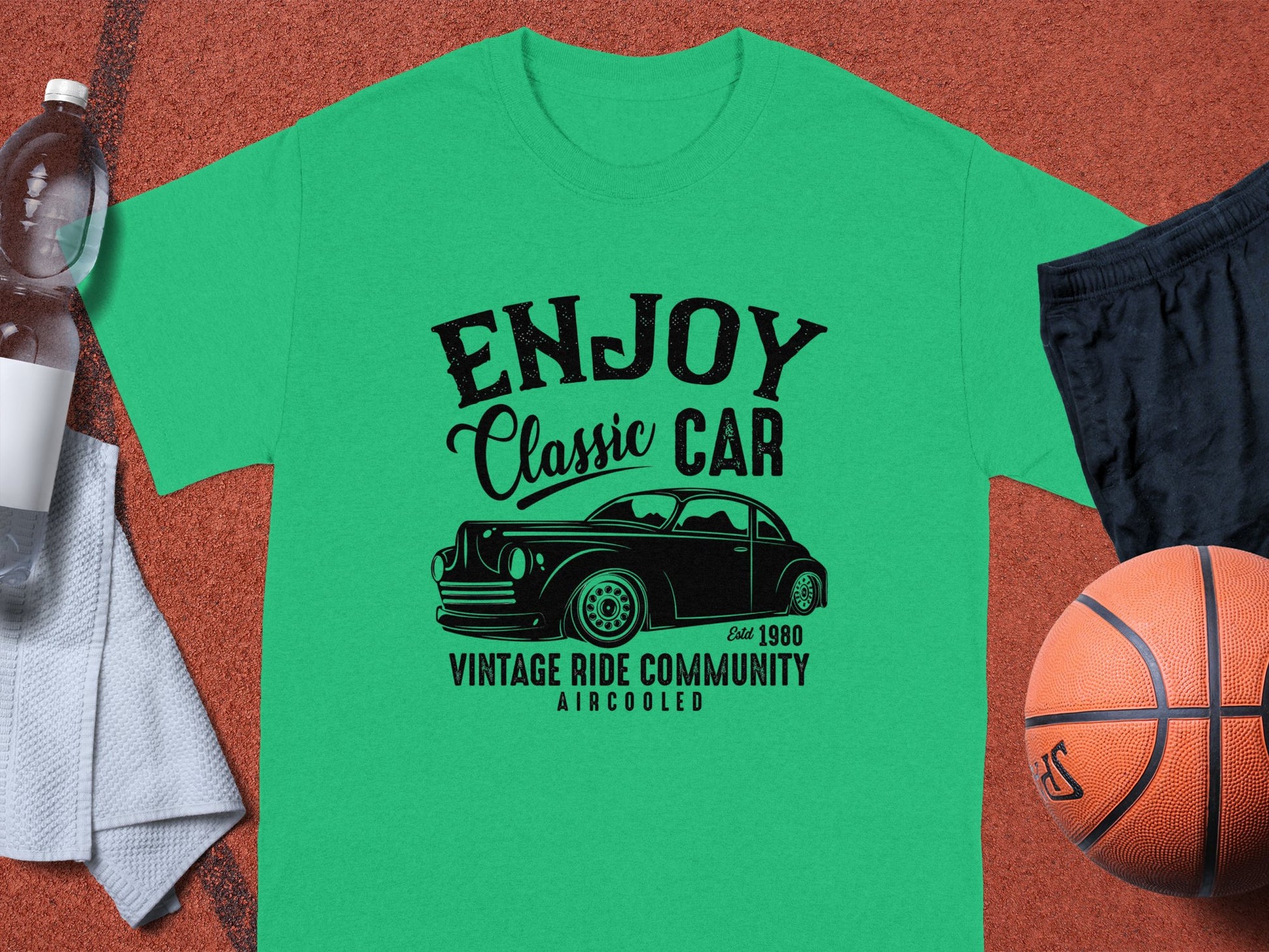 The Garment Graphics green t-shirt, ideal for car enthusiasts, showcases a Enjoy Classic Car design with a vintage car graphic and text Established 1980, Vintage Ride Community, Aircooled. Its displayed on a brown surface alongside a basketball, black shorts, towel, and water bottle.