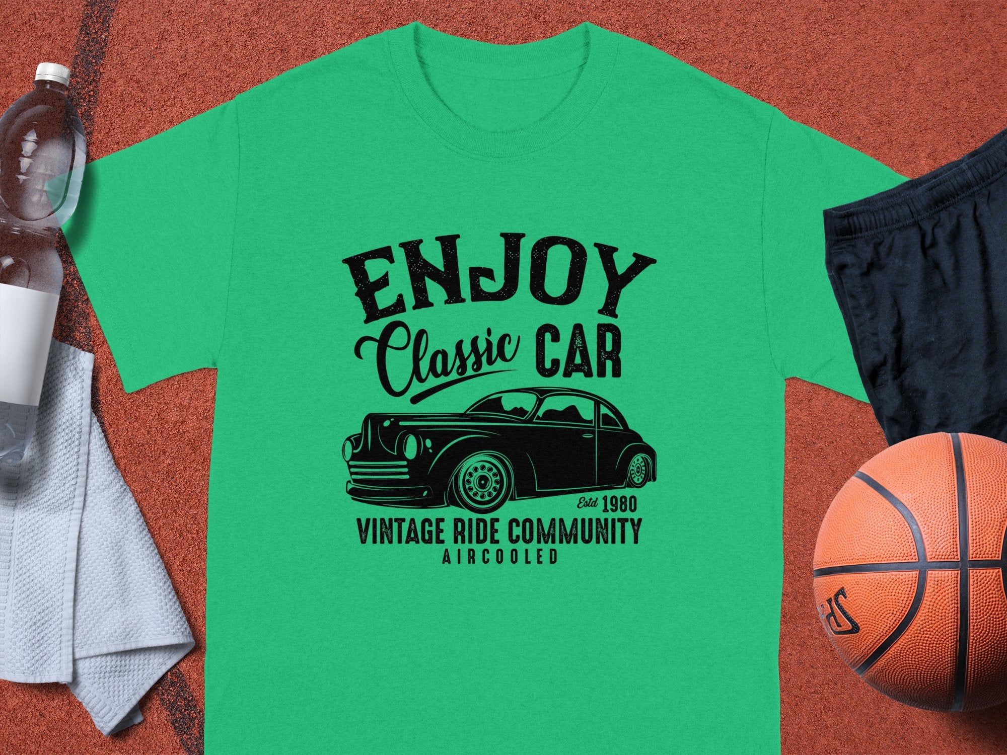 The Garment Graphics green t-shirt, ideal for car enthusiasts, showcases a Enjoy Classic Car design with a vintage car graphic and text Established 1980, Vintage Ride Community, Aircooled. Its displayed on a brown surface alongside a basketball, black shorts, towel, and water bottle.