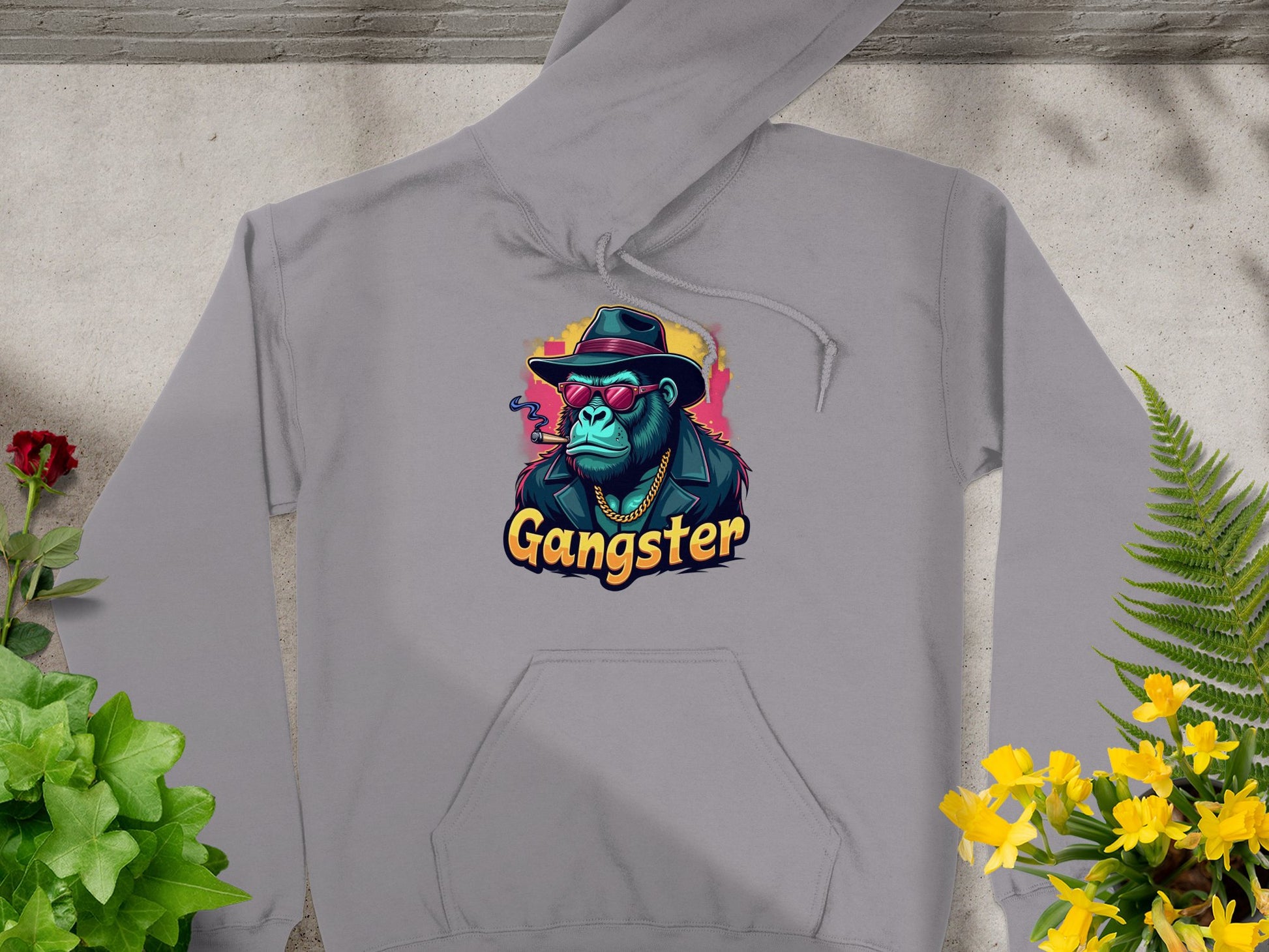 Garment Graphics presents a gray hipster urban hoodie featuring a graphic of a gangster ape in a hat, sunglasses, and chain, with Gangster in stylized text. Laid amidst green and yellow plants, its the epitome of trendy graphic hoodies.