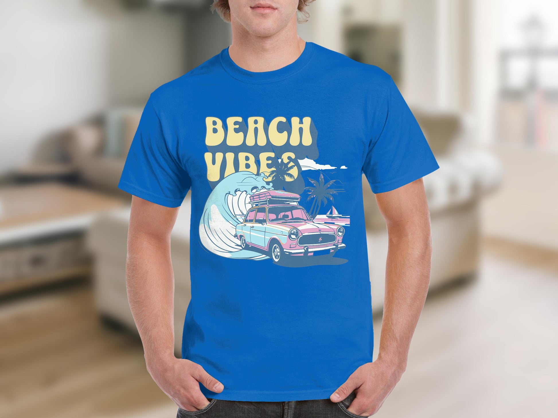 Someone wearing a Garment Graphics blue t-shirt with a Beach Vibe design that includes a classic car, surfboards, palm trees, and ocean waves. The setting is an indoor living room.
