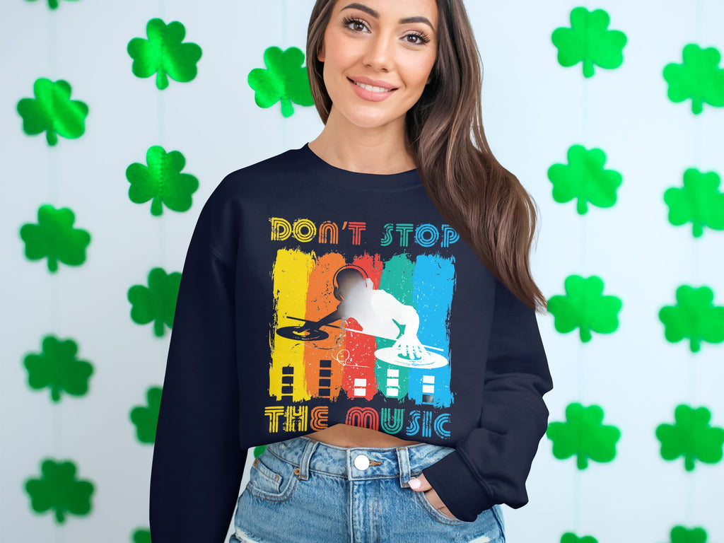 A woman smiles in front of a wall with green shamrocks, sporting a black Garment Graphics sweatshirt featuring a vibrant DJ design with turntables and the phrase Dont Stop the Music. Denim shorts complete her lively look. Perfect for music lovers!.