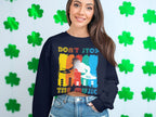 A woman smiles in front of a wall with green shamrocks, sporting a black Garment Graphics sweatshirt featuring a vibrant DJ design with turntables and the phrase Dont Stop the Music. Denim shorts complete her lively look. Perfect for music lovers!.