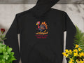 A Garment Graphics black hoodie with a Turkey On The Run graphic lies on concrete, surrounded by vibrant red, yellow, and green plants.