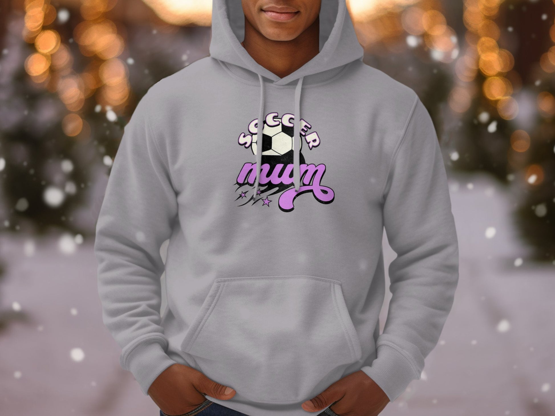 A person wears a cozy light gray hoodie from Garment Graphics, featuring a soccer ball with SOCCER MOM in white and pink lettering. This classic fit is perfect for any Soccer Mum, set against a bokeh lights background.