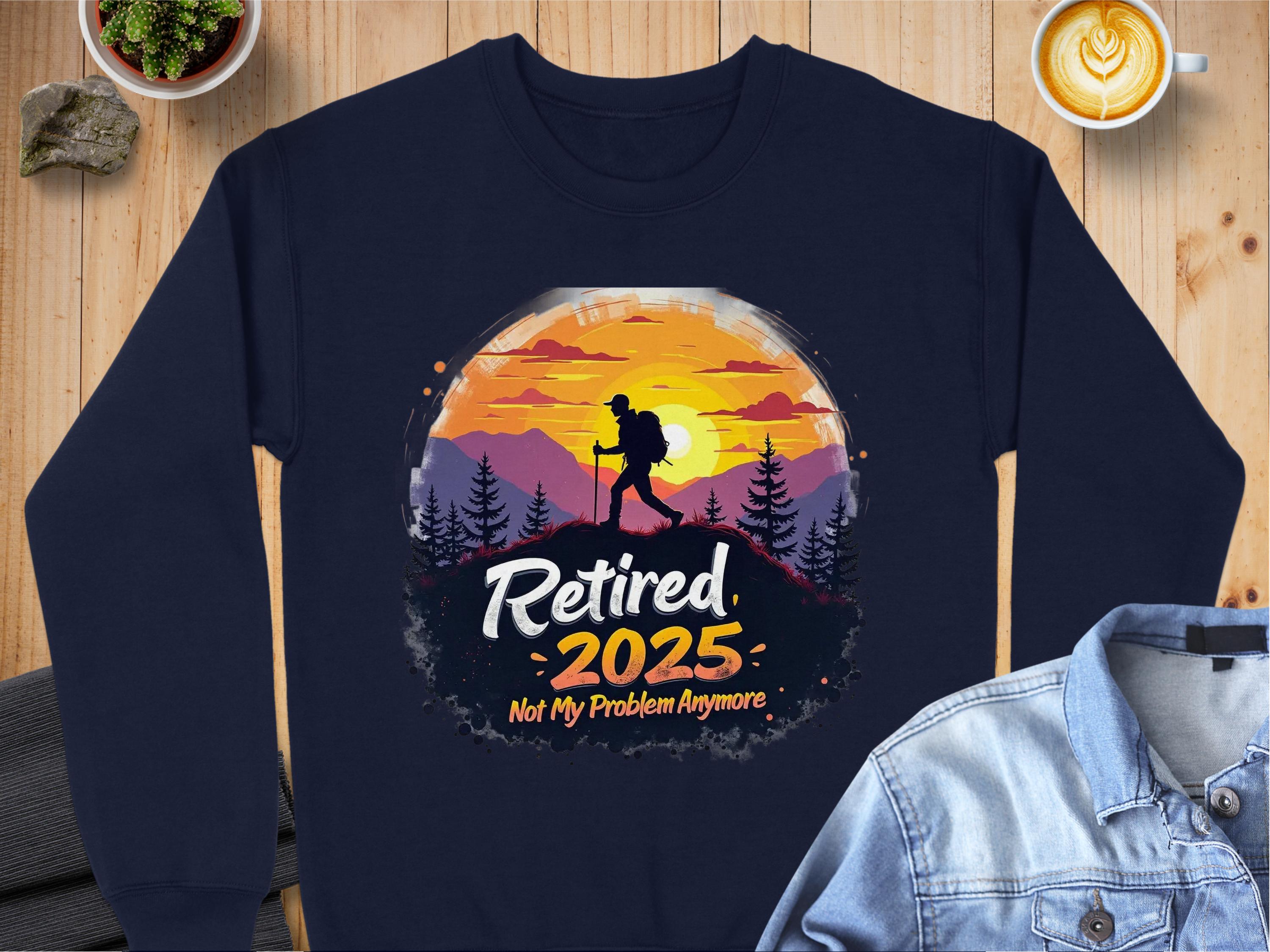 The Garment Graphics navy retirement sweatshirt shows a hiker silhouetted against a sunset and mountains, with Retired 2025: Not My Problem Anymore text. Available in unisex sizing, it’s displayed on a wooden surface next to a denim jacket, plants, and a latte.