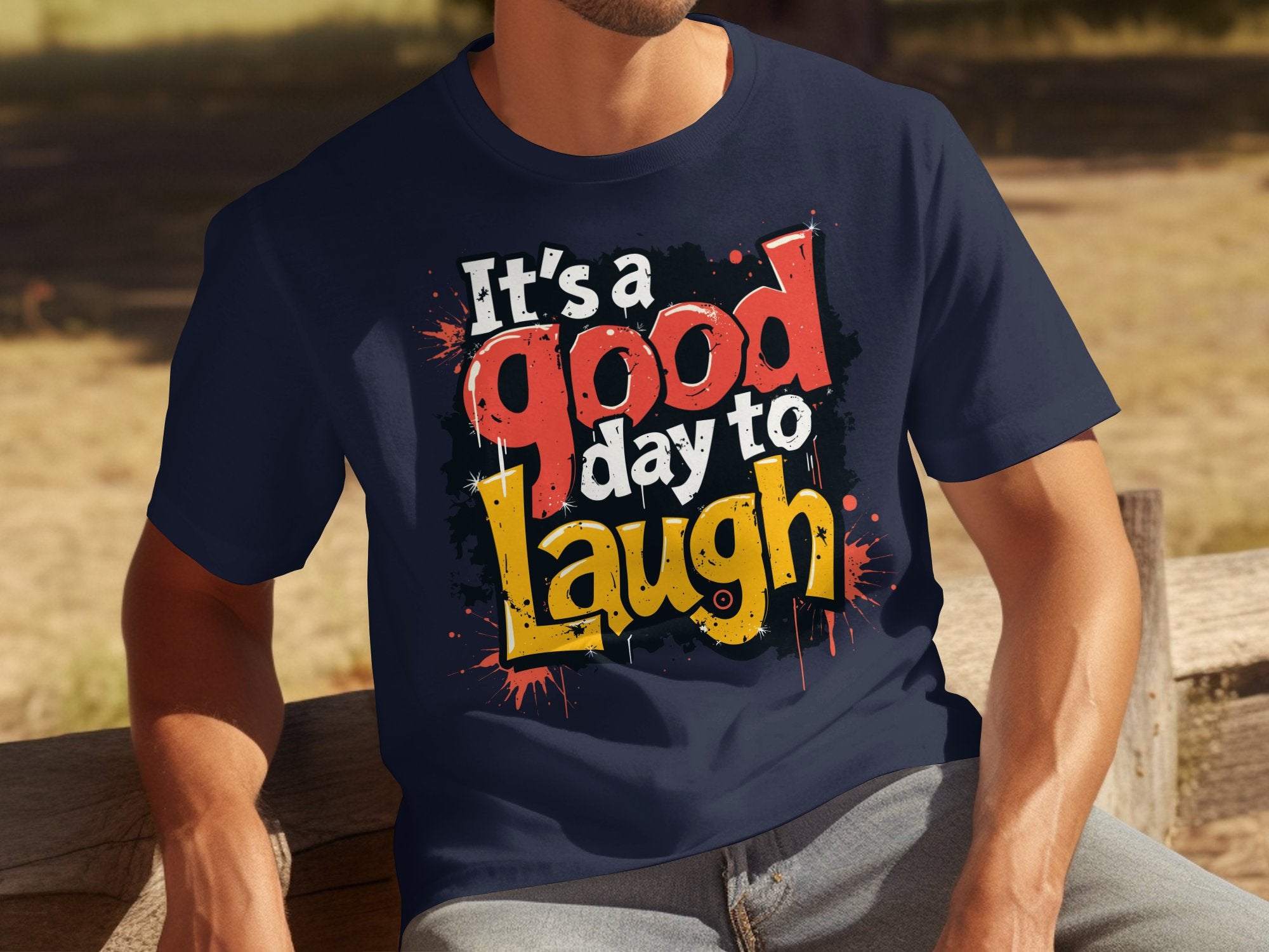 A person sits on a wooden bench wearing a navy blue Garment Graphics T-shirt with the message, Its a Good Day to Laugh, radiating vibrant colors against the blurred outdoor setting of grass and trees.