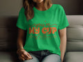 A person sits on a sofa wearing a 100% cotton green t-shirt from Garment Graphics that features Youre My Cup of Tea in brown lettering. With long hair cascading gently, they rest their arm on their knee, showcasing a delightful graphic tee.