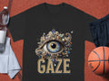A black T-shirt from Garment Graphics showcases an intricate eye illustration with GAZE beneath, elegantly laid on a red sports surface, paired with a basketball, black shorts, a water bottle, and a towel.