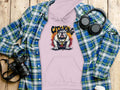 Casual look with a Garment Graphics light purple hoodie featuring a bulldog and City King text, matched with an urban blue-green plaid shirt. Nearby are a camera, headphones, and two drink containers on a wooden surface.