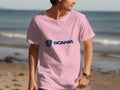 Dressed in a pink Scania logo T-shirt from Garment Graphics, a person stands by the shoreline. The calm ocean and sandy beach make a serene backdrop as they look right, their watch visible on their wrist.