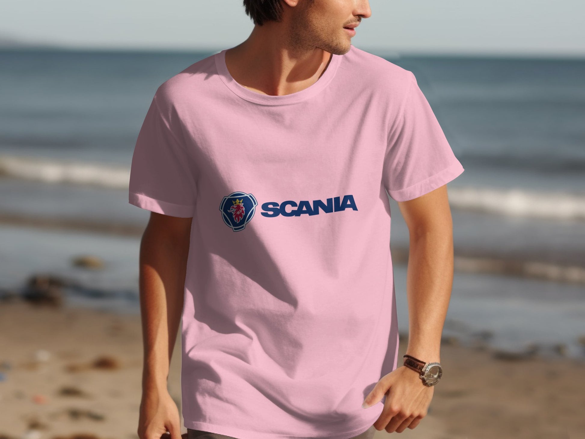 Dressed in a pink Scania logo T-shirt from Garment Graphics, a person stands by the shoreline. The calm ocean and sandy beach make a serene backdrop as they look right, their watch visible on their wrist.