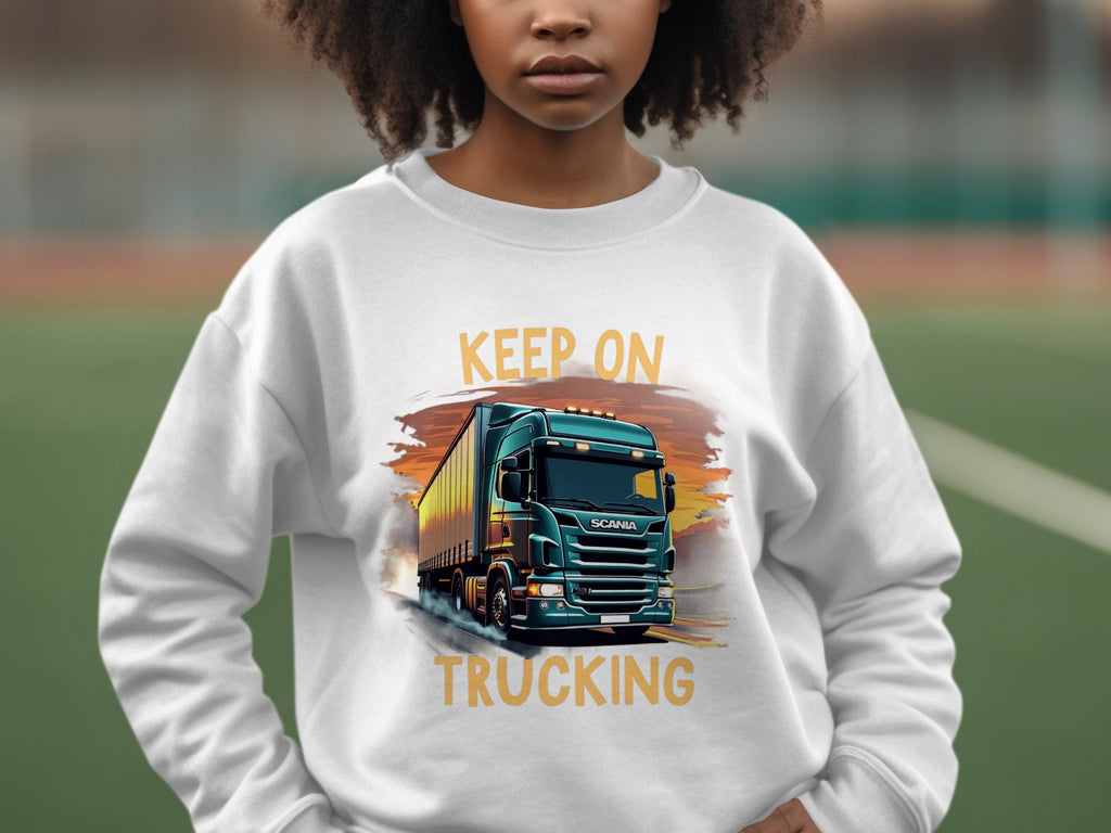 A truck enthusiast wears a Garment Graphics sweatshirt featuring a bold blue truck and the iconic KEEP ON TRUCKING text against the blurred backdrop of a sports field, enhancing the energetic vibe.