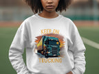 A truck enthusiast wears a Garment Graphics sweatshirt featuring a bold blue truck and the iconic KEEP ON TRUCKING text against the blurred backdrop of a sports field, enhancing the energetic vibe.