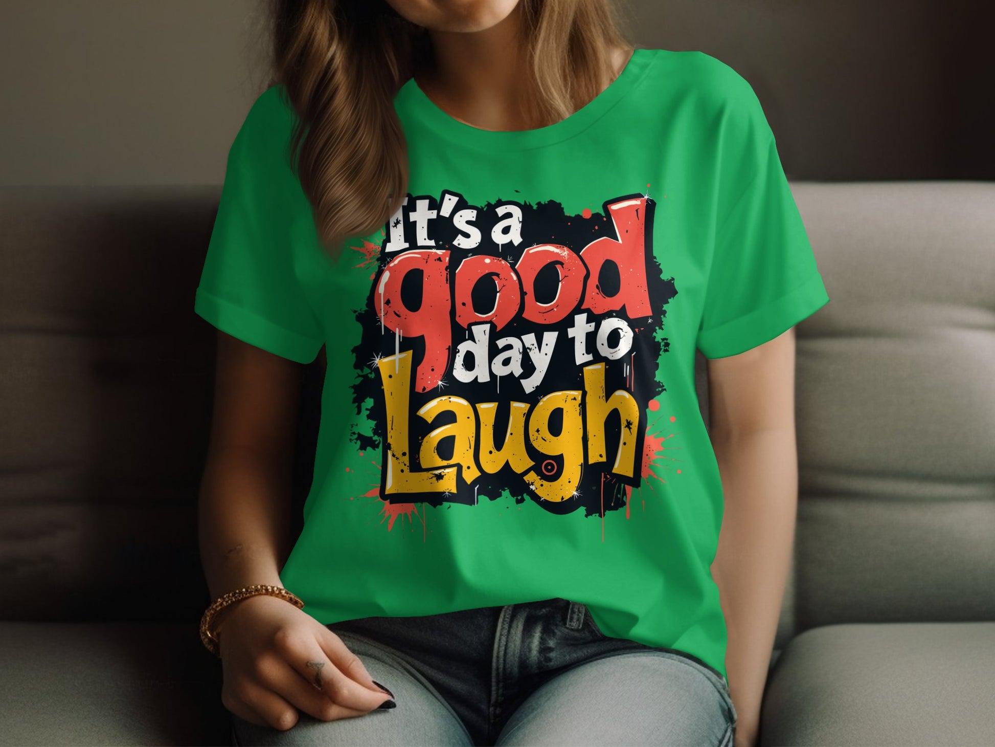A person with long hair, seen from the shoulders down, is on a couch wearing Garment Graphics semi-fitted green T-shirt. It has Its a good day to laugh in bold, colorful lettering and a playful paint splatter effect that exudes positivity.