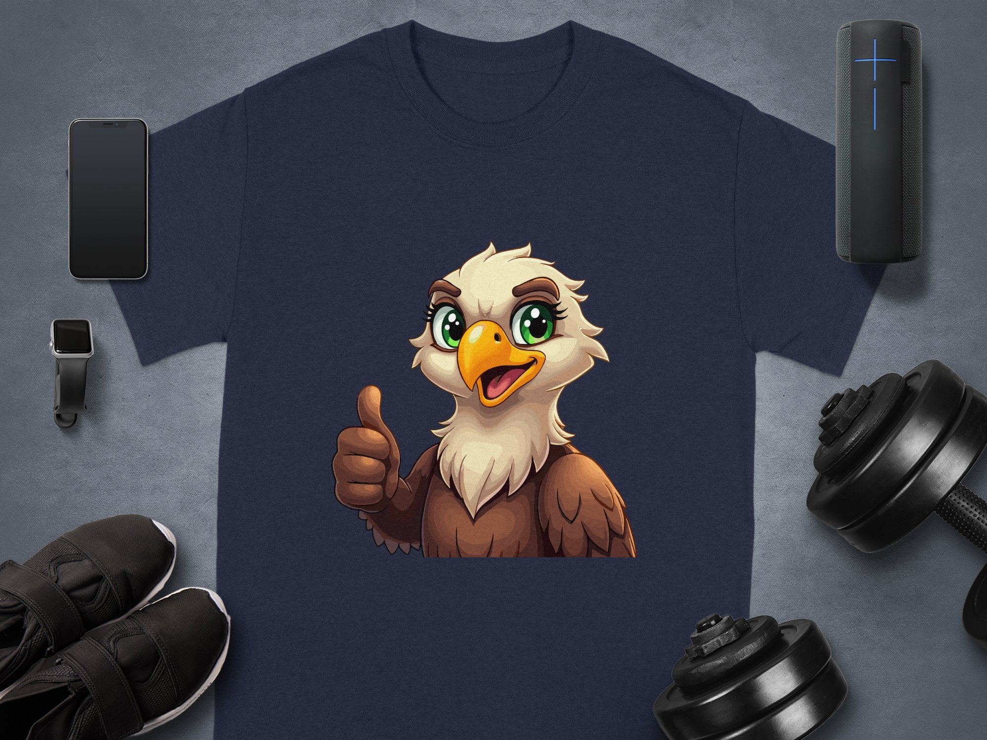 The Garment Graphics navy blue Eagle T-shirt has a cartoon eagle giving a thumbs up. Ideal for your casual wardrobe, its showcased with a smartphone, wireless speaker, smartwatch, black sneakers, and adjustable dumbbells on a gray background.