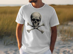 Wearing a daring white graphic tee with a skull and crossbones design by Garment Graphics, a person stands in sandy terrain surrounded by tall grass against a blurred backdrop.
