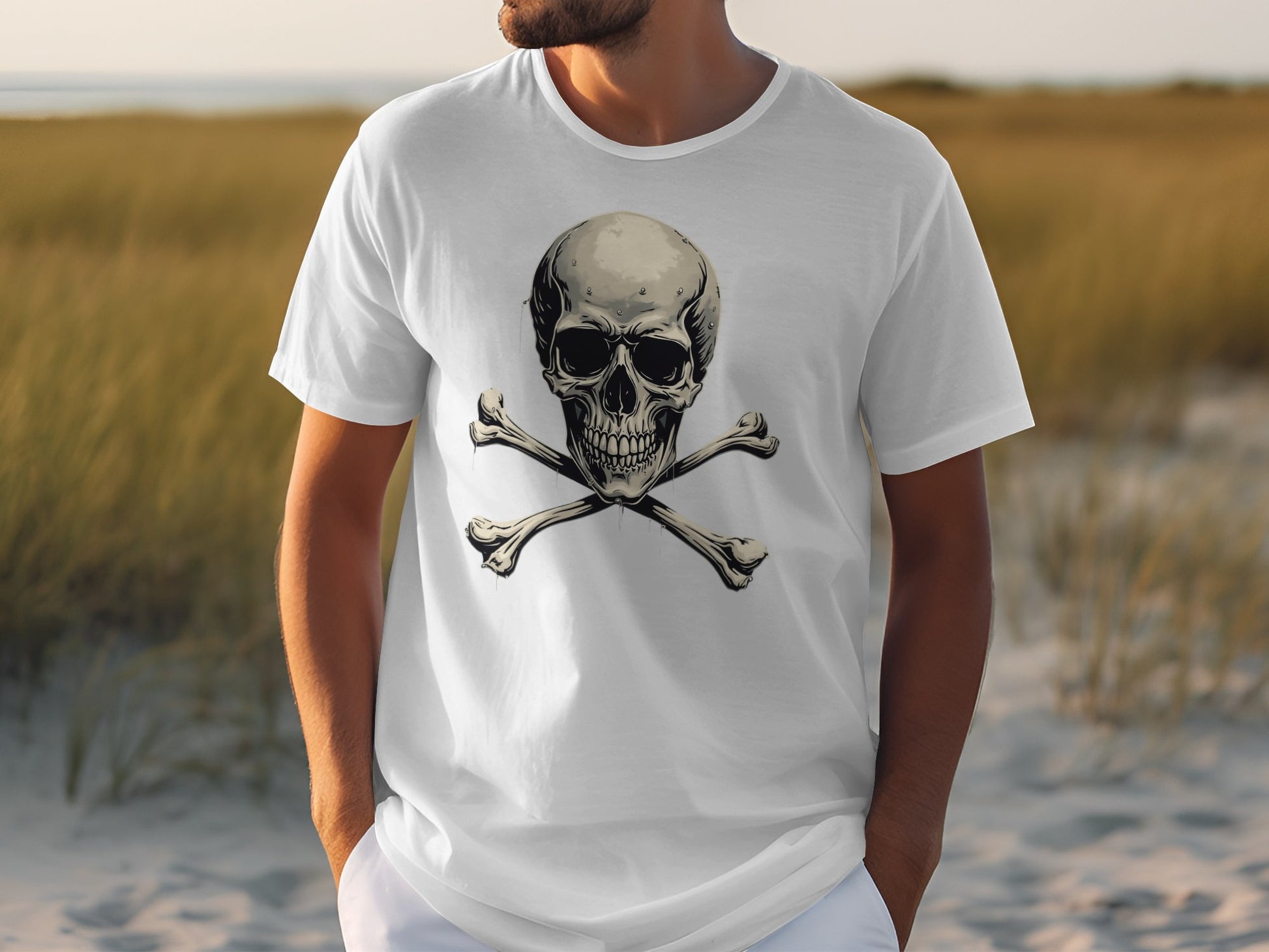 Wearing a daring white graphic tee with a skull and crossbones design by Garment Graphics, a person stands in sandy terrain surrounded by tall grass against a blurred backdrop.