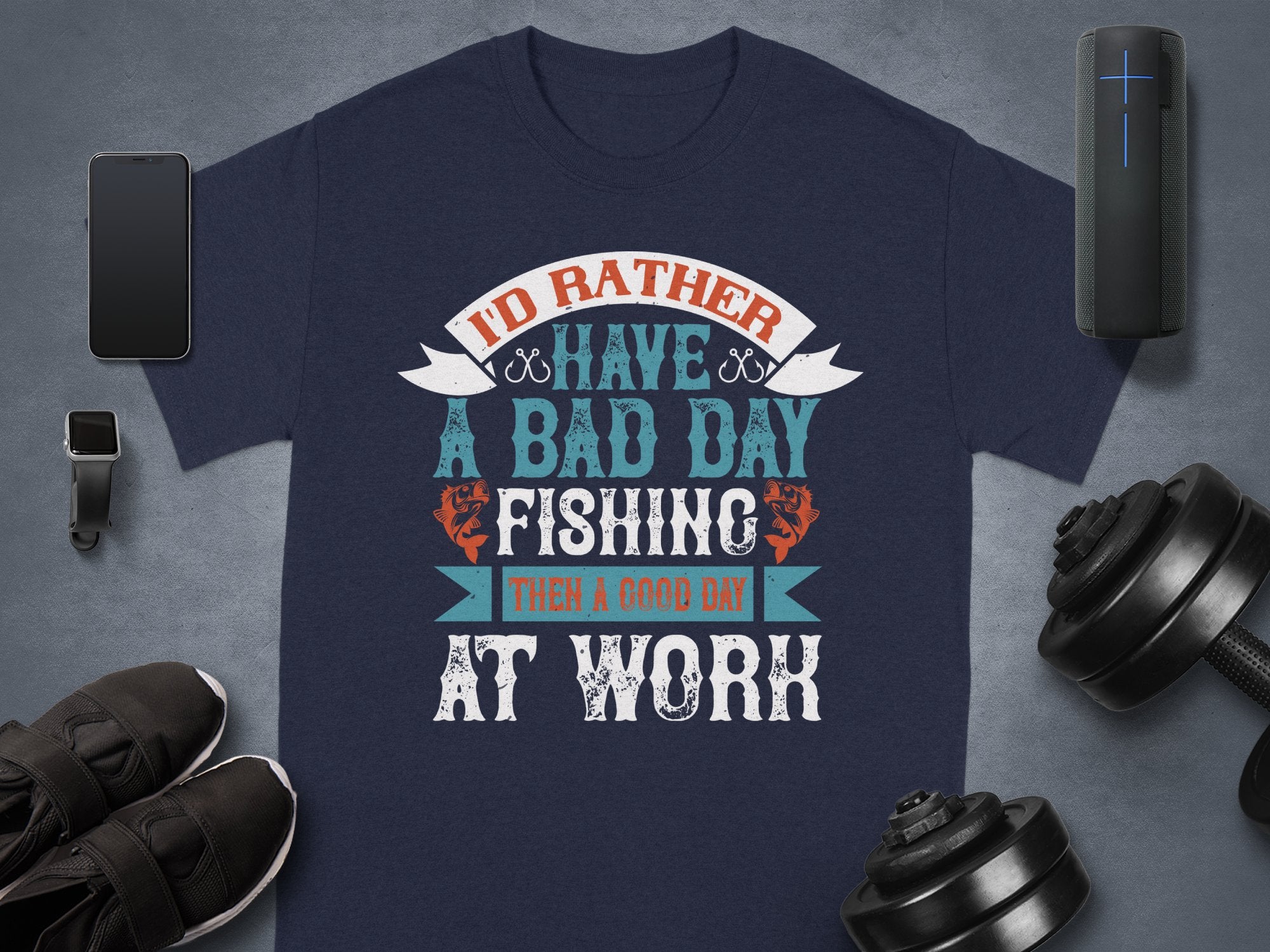A Garment Graphics navy blue fishing T-shirt with Id rather have a bad day fishing than a good day at work is surrounded by a smartphone, smartwatch, wireless speaker, shoes, and dumbbells on a gray background. Its an ideal gift for anglers who cherish relaxed days by the water.
