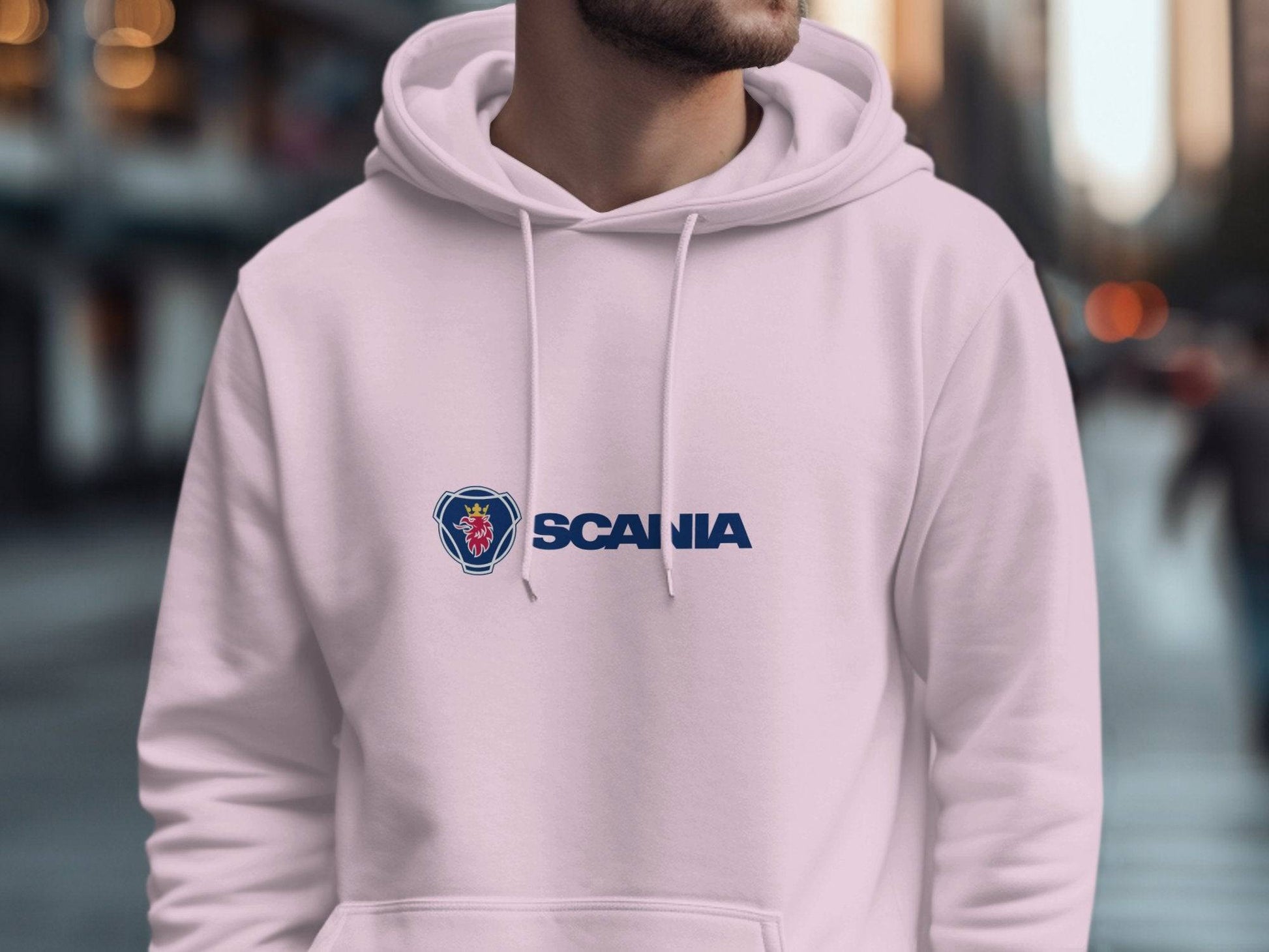 A person wearing a light-colored Garment Graphics hoodie with the iconic Scania logo stands in an urban setting. The blurred background of buildings and streets makes this unique fashion piece stand out vividly.