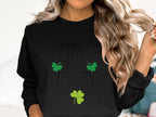 A person with wavy blonde hair wears a Garment Graphics black sweatshirt, reading Happy St. Patricks Day, featuring three green shamrocks and two overlapping hearts, complementing their carefree gesture as they touch their hair.