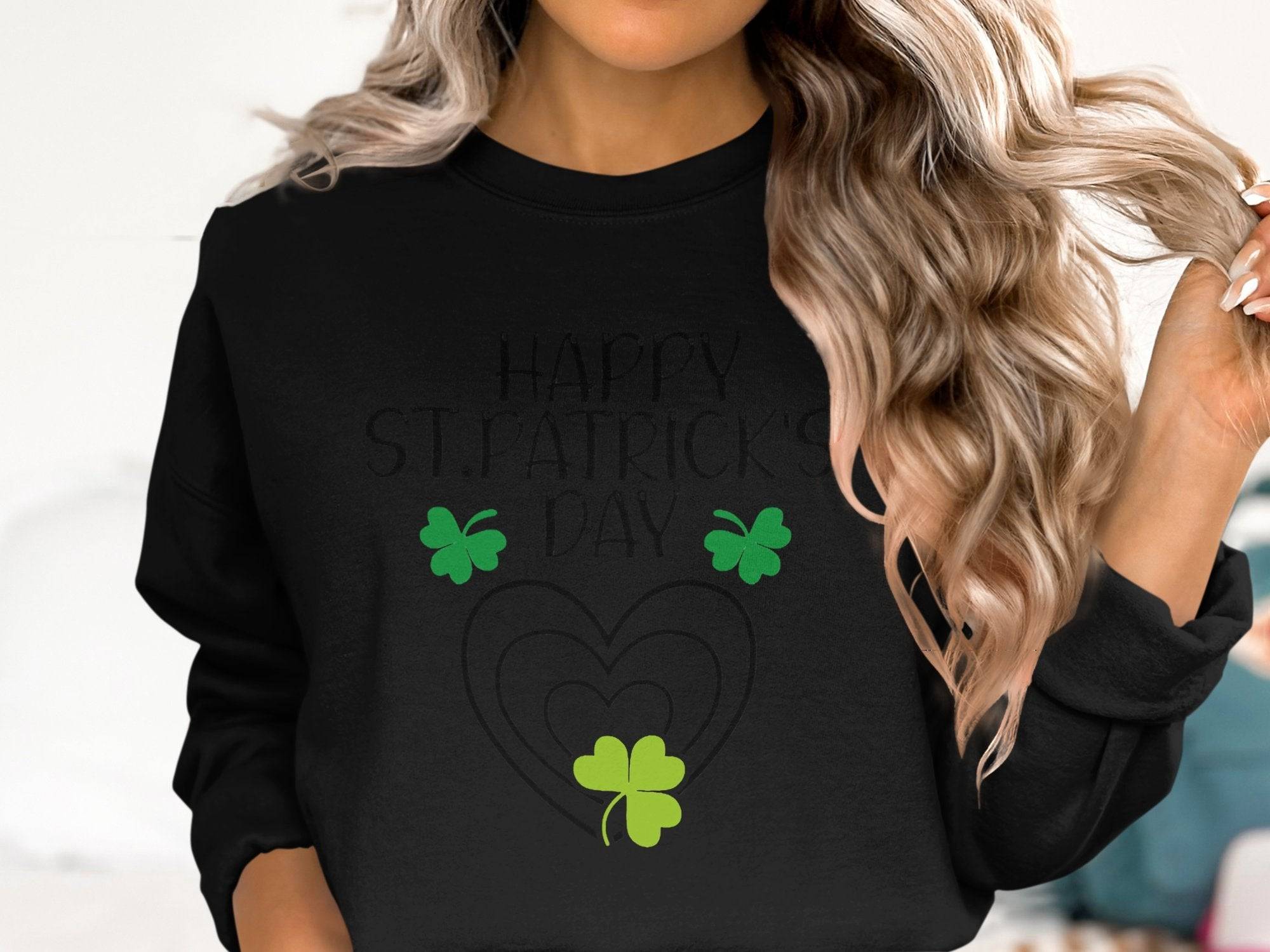 A person with wavy blonde hair wears a Garment Graphics black sweatshirt, reading Happy St. Patricks Day, featuring three green shamrocks and two overlapping hearts, complementing their carefree gesture as they touch their hair.