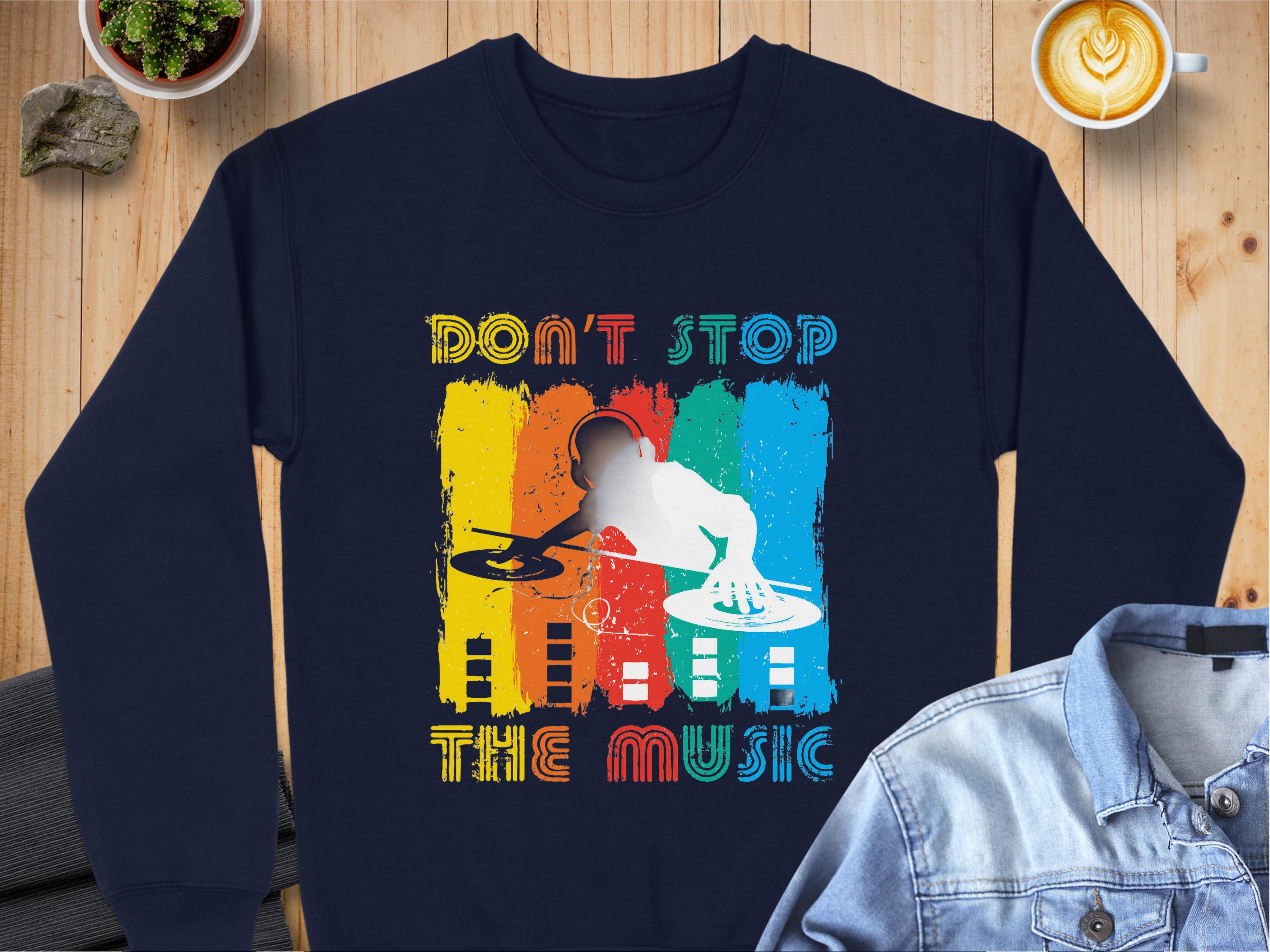 A navy sweatshirt from Garment Graphics lies on a wooden table, featuring a DJ design with vibrant bars and the words Dont Stop the Music. Nearby are a denim jacket, coffee, and a plant.