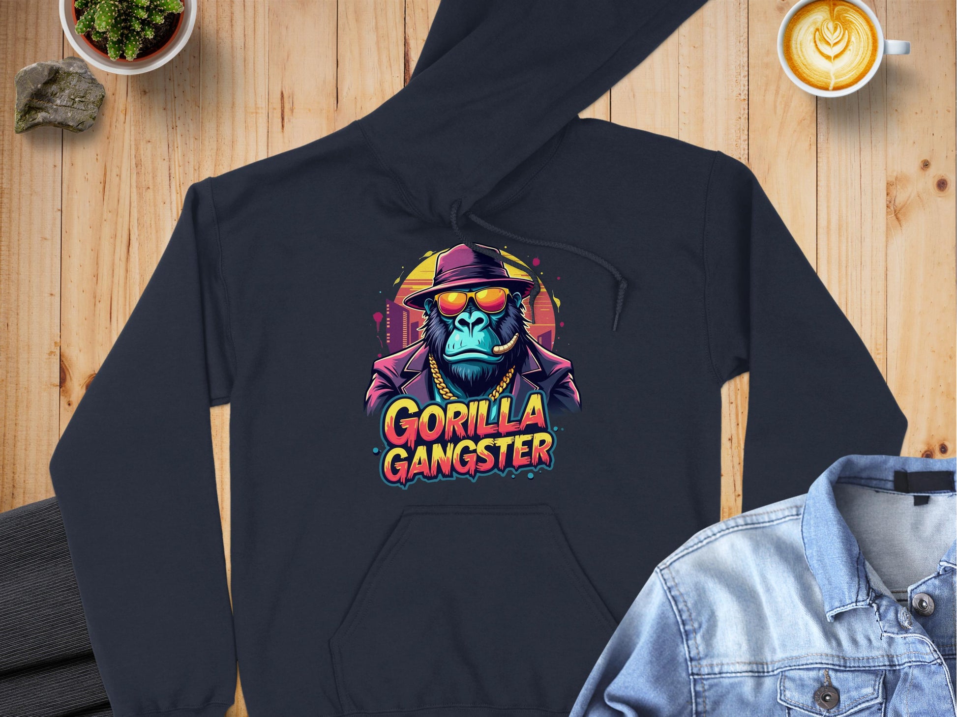 The Garment Graphics hoodie showcases a gorilla in a hat and sunglasses, labeled Gorilla Gangster. Placed on wood with a denim jacket, small plant, latte, and rock nearby, this streetwear piece combines style with bold gorilla art.