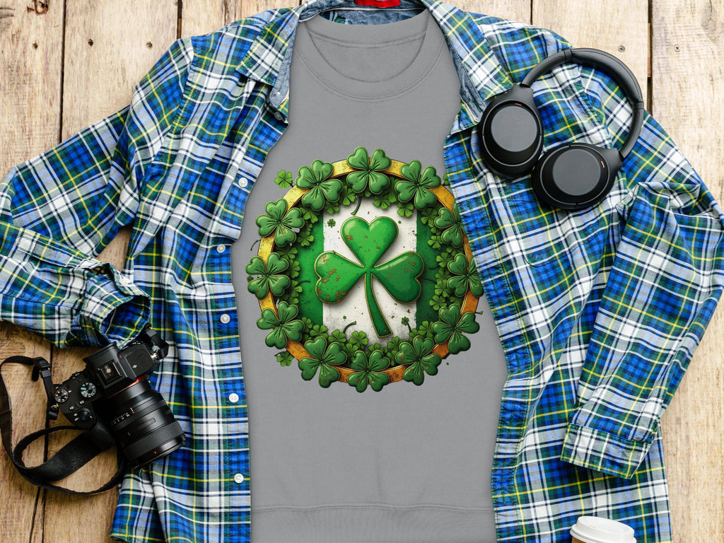 A Garment Graphics gray t-shirt with a lucky four-leaf clover design lies on a wooden surface, topped by a blue and green plaid shirt embodying the Irish spirit. Nearby rest black headphones, a camera, and a white coffee cup.