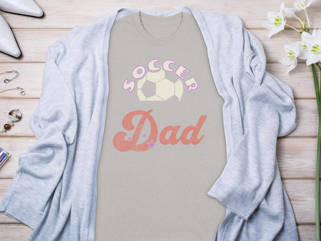 A Garment Graphics light gray Soccer Dad T-shirt with a soccer ball design is laid flat, perfectly paired with a cozy cardigan, white shoes, assorted jewelry, and a small plant on the wooden surface.