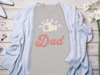 A Garment Graphics light gray Soccer Dad T-shirt with a soccer ball design is laid flat, perfectly paired with a cozy cardigan, white shoes, assorted jewelry, and a small plant on the wooden surface.