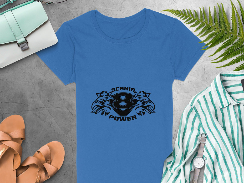 A blue T-shirt by Garment Graphics features the SCANIA 8 POWER logo surrounded by lions, crowns, and eagle heads. It sits on a gray background next to a green-striped T-shirt, tan sandals, a light blue purse, and fern leaves.