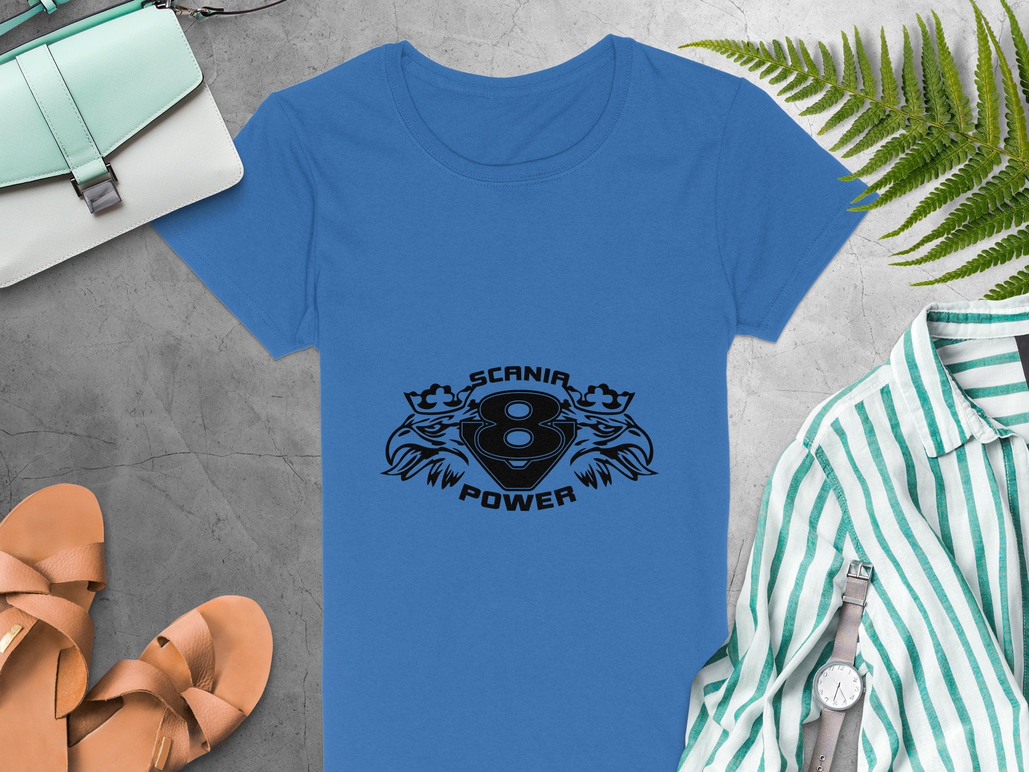 A blue T-shirt by Garment Graphics features the SCANIA 8 POWER logo surrounded by lions, crowns, and eagle heads. It sits on a gray background next to a green-striped T-shirt, tan sandals, a light blue purse, and fern leaves.