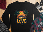 A Garment Graphics black sweatshirt features a One Love Sunset scene with palm trees, a sunset, and an airplane, finished with bold One Love text. It pairs perfectly on a wooden surface with accessories like a red knit hat, gloves, and a plaid shirt and scarf.