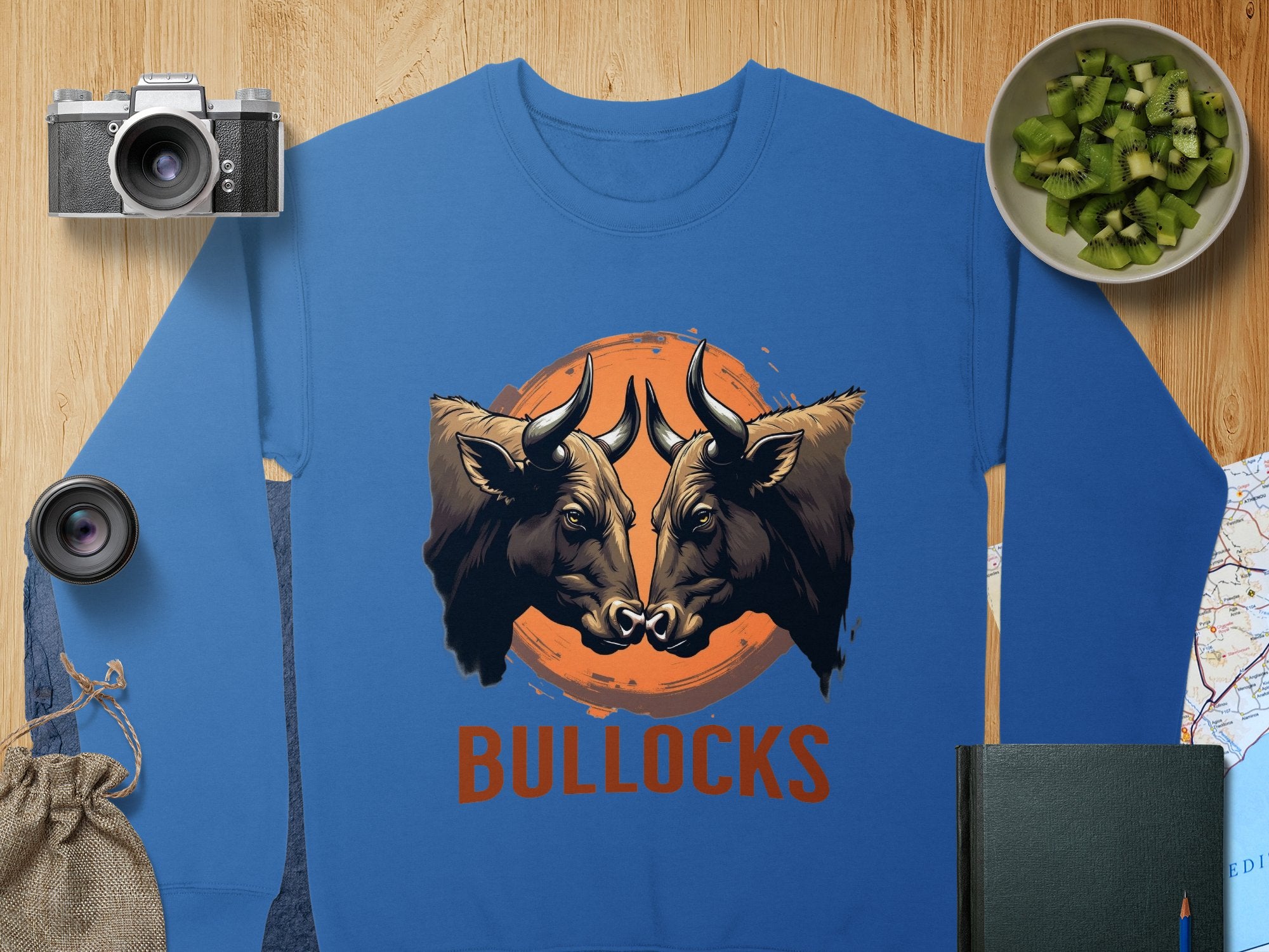 Garment Graphics blue sweatshirt showcases a striking bullocks print with two bulls facing an orange circle and BULLOCKS below. It is displayed among a camera, lens, cucumbers in a bowl, map, book, and small bag on a wooden surface.