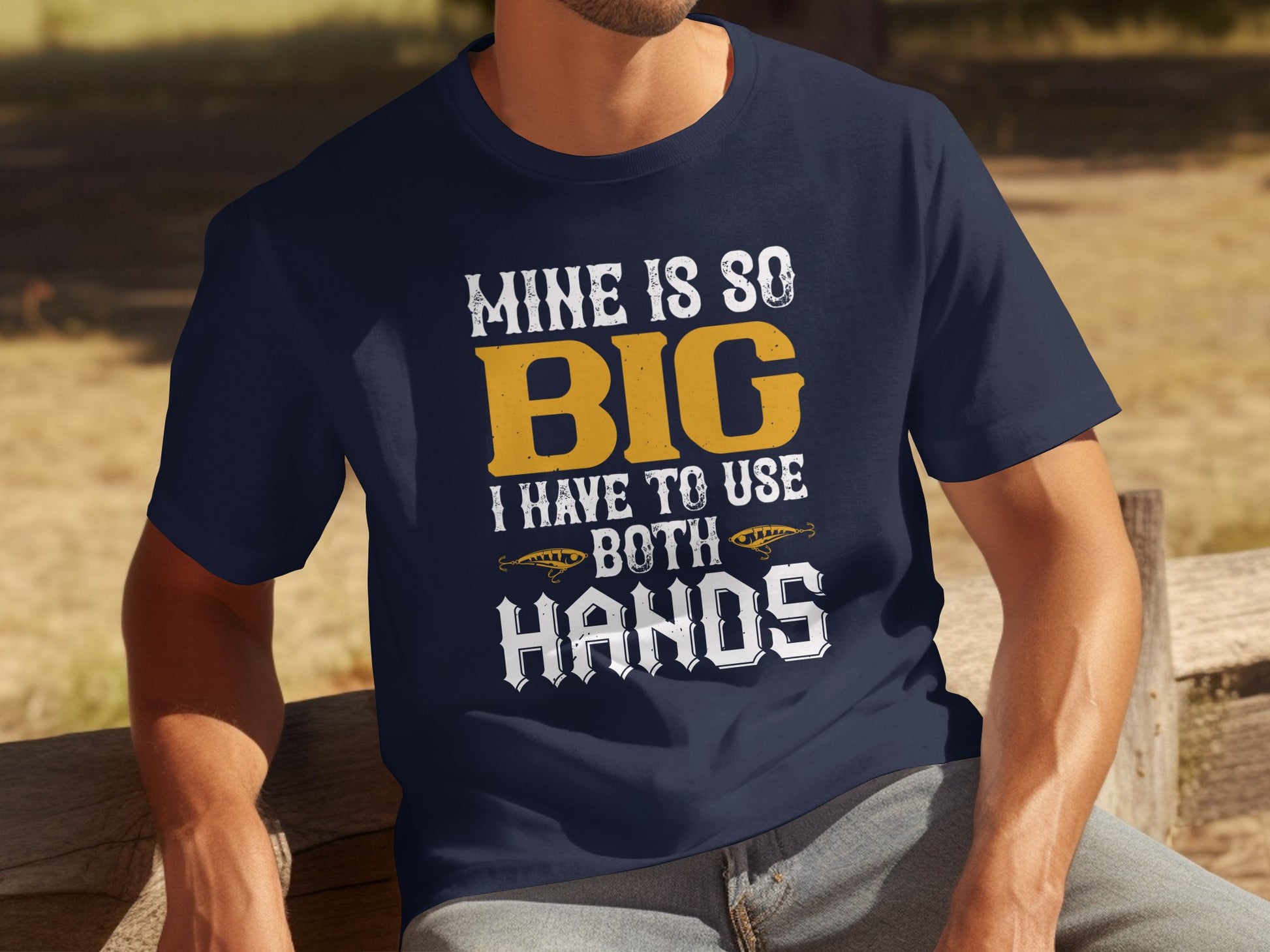 A person relaxes on an outdoor wooden bench in a navy blue Garment Graphics tee with bold yellow and white text reading, Mine is so big I have to use both hands, featuring playful football graphics.