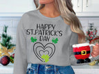 A person with long hair holds a Santa-themed mug and wears a Garment Graphics festive sweatshirt with Happy St. Patricks Day and clover designs, standing in a kitchen decorated festively with white cabinets.
