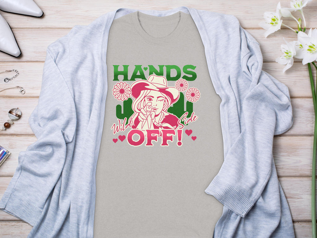 A light grey t-shirt from Garment Graphics showcases a bold design of a person in a hat making a peace sign. The text reads, HANDS OFF! Wild & Free. Its laid on wood, surrounded by a cardigan, shoes, flowers, and jewelry.