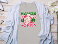A light grey t-shirt from Garment Graphics showcases a bold design of a person in a hat making a peace sign. The text reads, HANDS OFF! Wild & Free. Its laid on wood, surrounded by a cardigan, shoes, flowers, and jewelry.