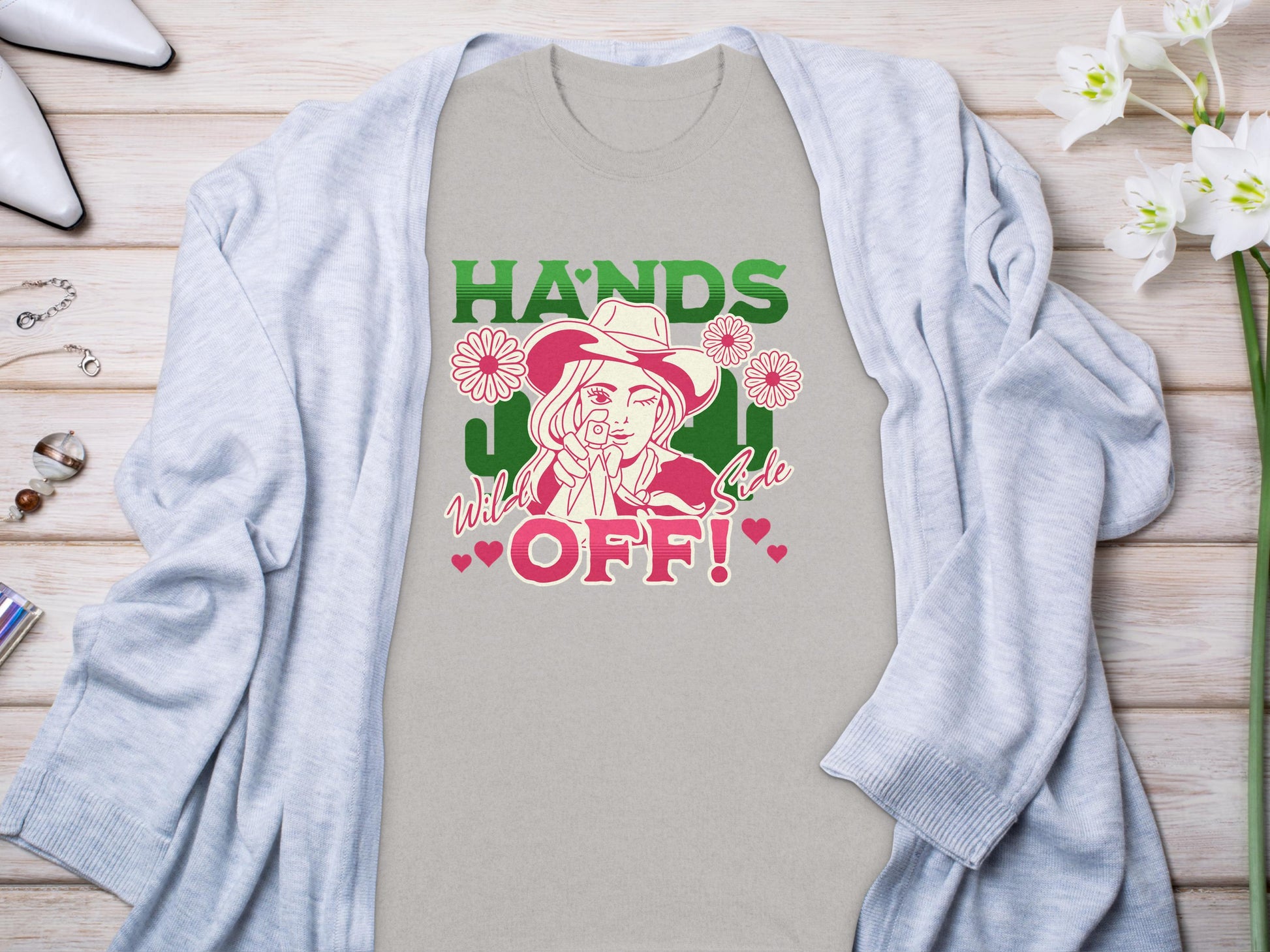 A light grey t-shirt from Garment Graphics showcases a bold design of a person in a hat making a peace sign. The text reads, HANDS OFF! Wild & Free. Its laid on wood, surrounded by a cardigan, shoes, flowers, and jewelry.