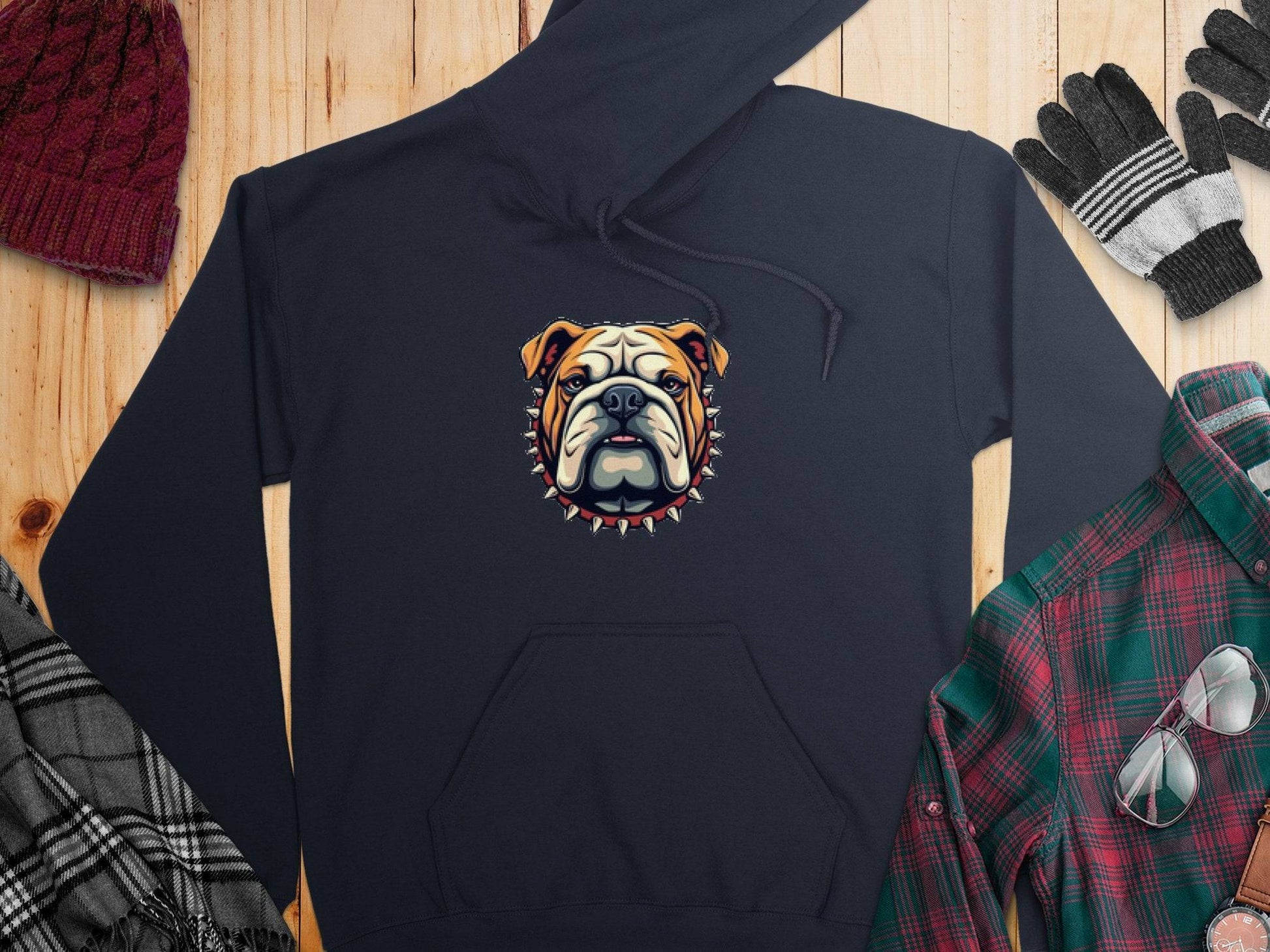 A black bulldog hoodie with a spiked collar design by Garment Graphics lies on a wooden surface. Surrounding it are a maroon knit hat, striped gloves, a gray plaid scarf, and a green and red plaid shirt, making a bold statement in cozy accessories.