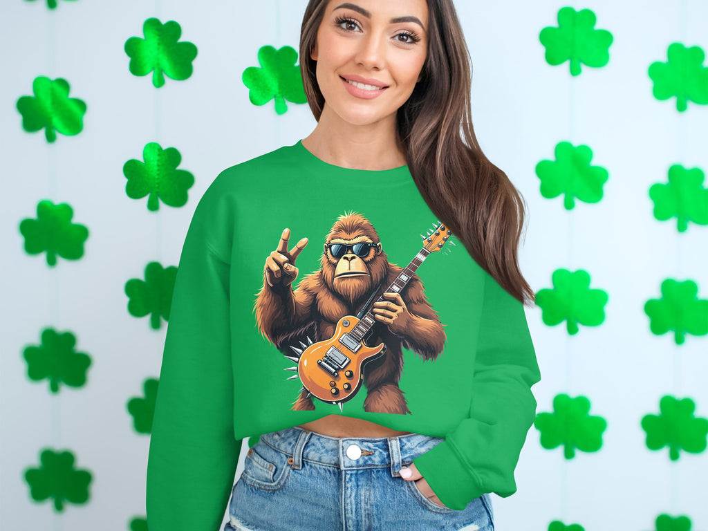 A smiling woman with long brown hair wears denim shorts and a Garment Graphics green sweatshirt featuring a cartoon Bigfoot playing an electric guitar with a peace sign, set against a shamrock pattern. .