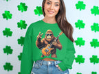 A smiling woman with long brown hair wears denim shorts and a Garment Graphics green sweatshirt featuring a cartoon Bigfoot playing an electric guitar with a peace sign, set against a shamrock pattern. .