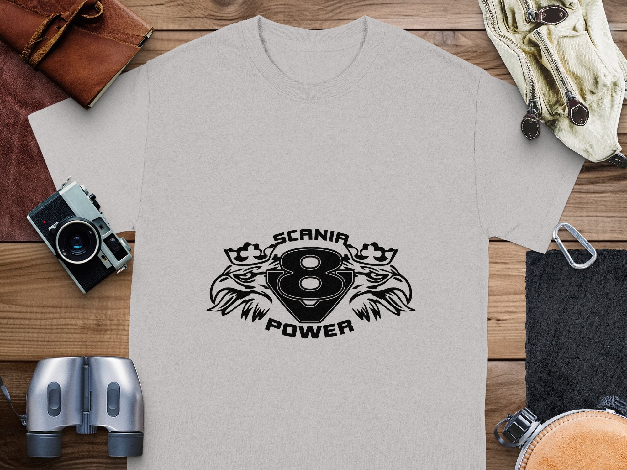 A gray T-shirt by Garment Graphics lies on a wooden surface, featuring the SCANIA POWER EAGLE graphic with an 8 between wings and crowns. Nearby are a leather pouch, gloves, camera, binoculars, and a black notebook—ideal for the adventurous spirit.