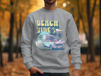 A person wears a Garment Graphics unisex graphic sweatshirt depicting a vintage car with a surfboard, palm trees, and waves, capturing retro roadtrip allure. Beach Vibes is printed boldly against an autumn tree-lined path backdrop, ensuring universal fit and style.