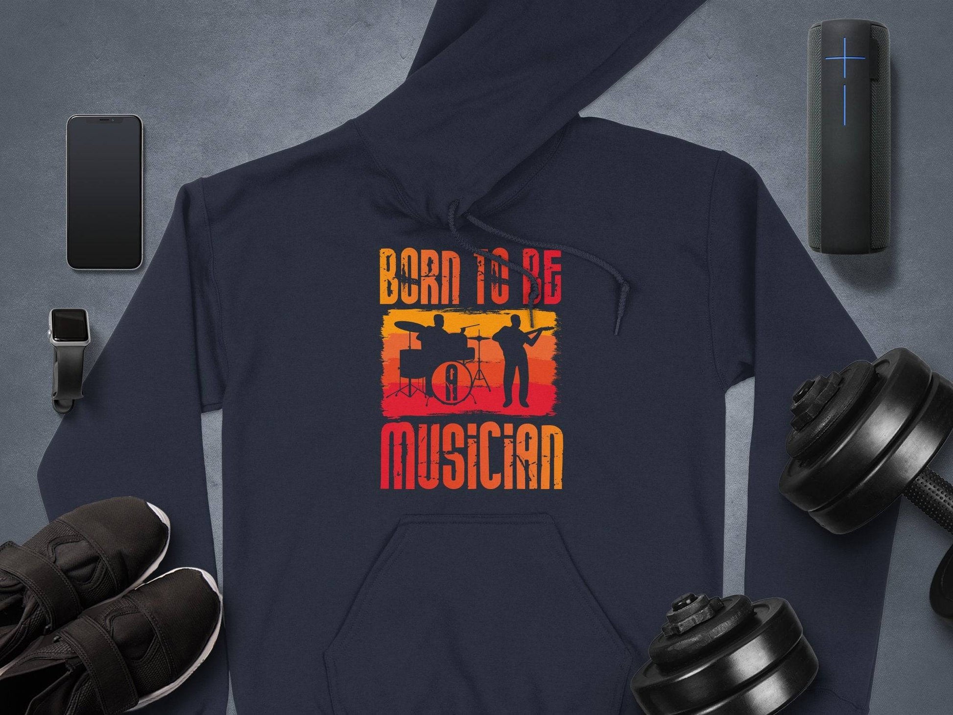 Garment Graphics dark unisex fashion hoodie features a drummer and guitarist silhouette with Born to be Musician in vibrant orange. Ideal for music enthusiasts, its displayed on a sleek gray surface beside a smartphone, smartwatch, wireless speaker, athletic shoes, and dumbbells.