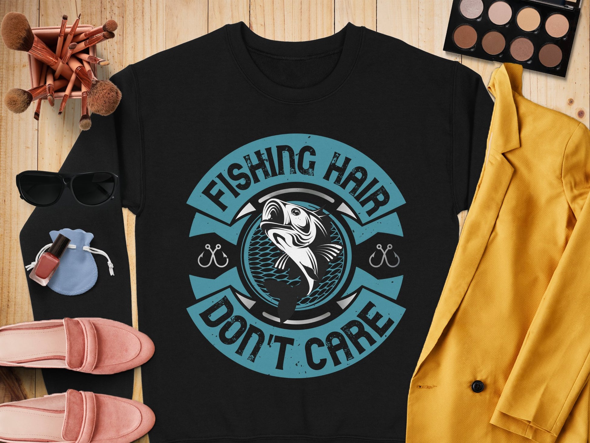 The Garment Graphics black shirt features a circular fish logo and the text, Fishing Hair Dont Care. Its stylishly paired with a mustard jacket, makeup palette, sunglasses, red shoes, brushes, and a pouch on a wooden surface—ideal for fishing enthusiasts.