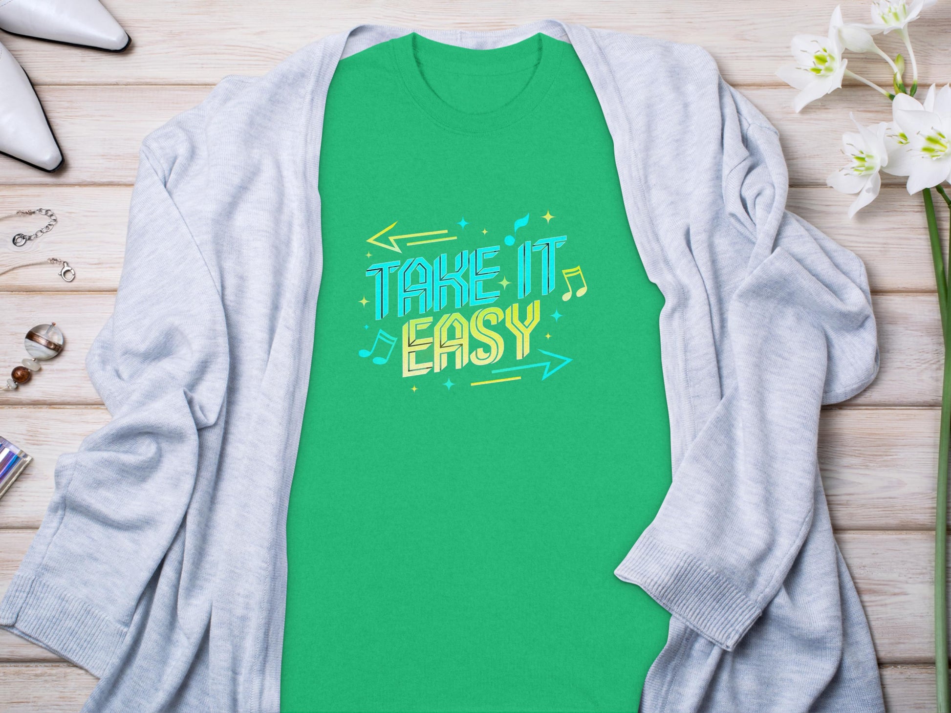 A vibrant Garment Graphics green Take It Easy T-Shirt with colorful letters, musical notes, and arrows lies flat on a wooden surface under a light gray cardigan, surrounded by white flowers, jewelry, and high-heeled shoes.